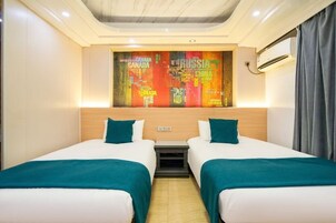 Habitación de hotel con dos camas, ropa de cama color aguamarina, cabecero de madera, unidad de aire acondicionado montada en la pared y una obra de arte abstracta y colorida sobre las camas.