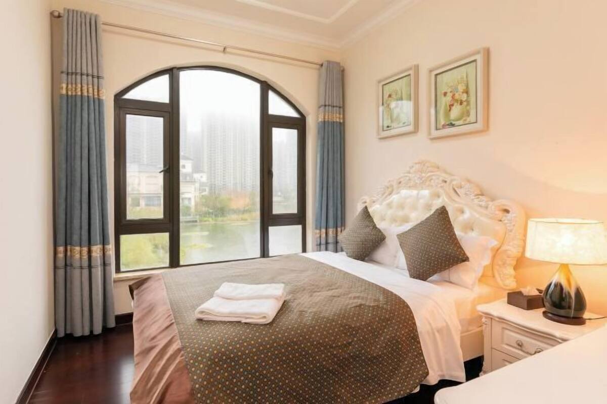 A hotel room with a large arched window, a bed with a white headboard, two framed pictures on the wall, and a bedside lamp.