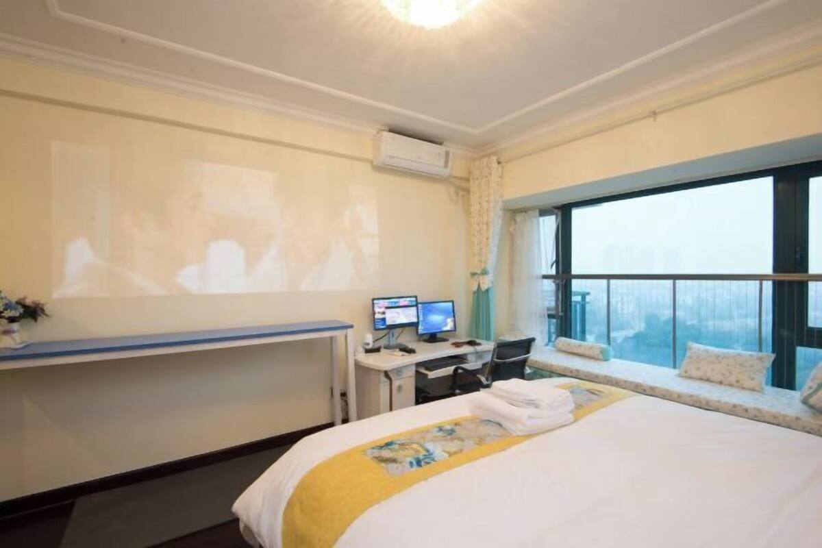 A hotel room with a bed, a desk with two monitors, a window with a city view, and an air conditioning unit.