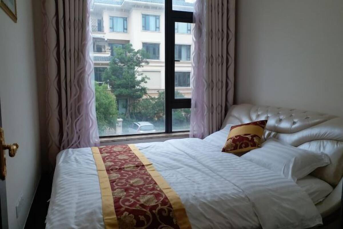 A bedroom with a large window, a bed with a white and red bedspread, and a view of buildings outside.