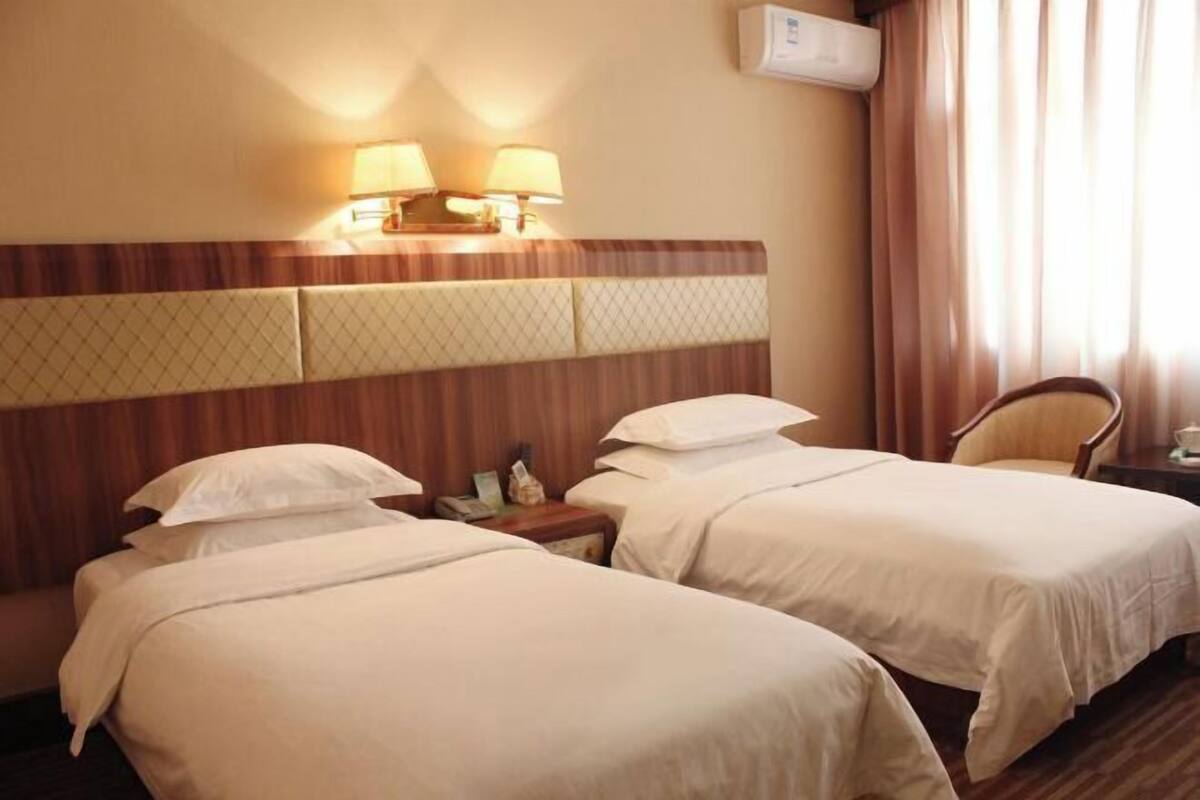 A hotel room with two beds, a wooden headboard, bedside lamps, a chair, and an air conditioning unit.