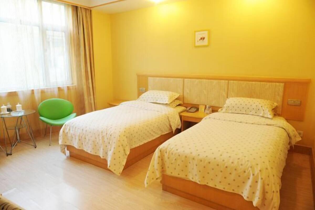 A hotel room with two beds, a small table, a green chair, and a window with curtains.
