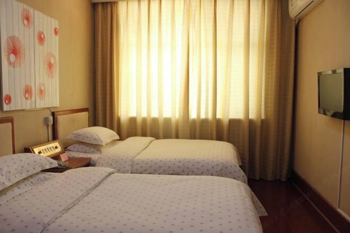 A hotel room with two beds, a bedside table with a lamp, a wall-mounted TV, and a window with curtains.