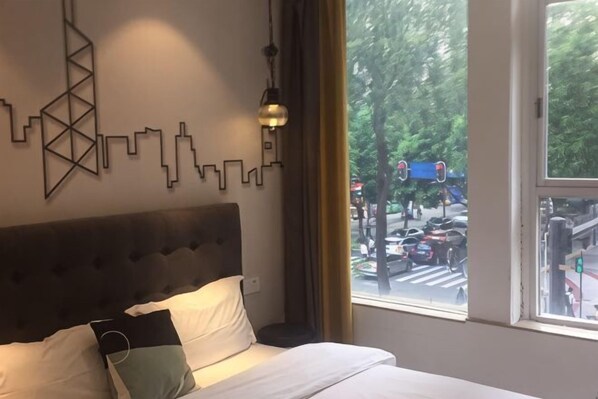 A hotel room with a bed, a cityscape wall art, a window with a view of the street, and a hanging light fixture.