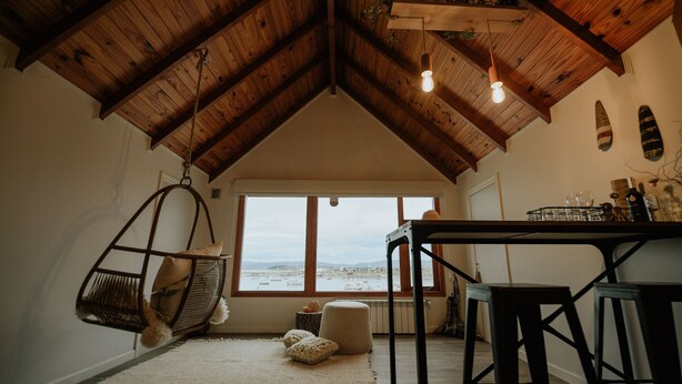 A cozy room with a high wooden ceiling, a hanging chair, a table with stools, and a view of the sea through large windows.