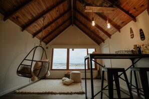 Una stanza accogliente con soffitto in legno a volta, una poltrona sospesa, un tavolo con sgabelli e vista sul mare attraverso ampie finestre.