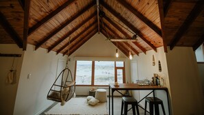 A cozy room with a sloped wooden ceiling, a hanging chair, a small round table, and a view of a landscape through large windows.