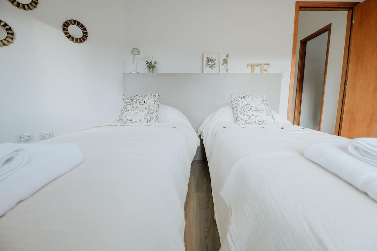 Une chambre double avec deux lits, une table de chevet avec une lampe, une petite étagère avec des objets décoratifs et une porte sur la droite.