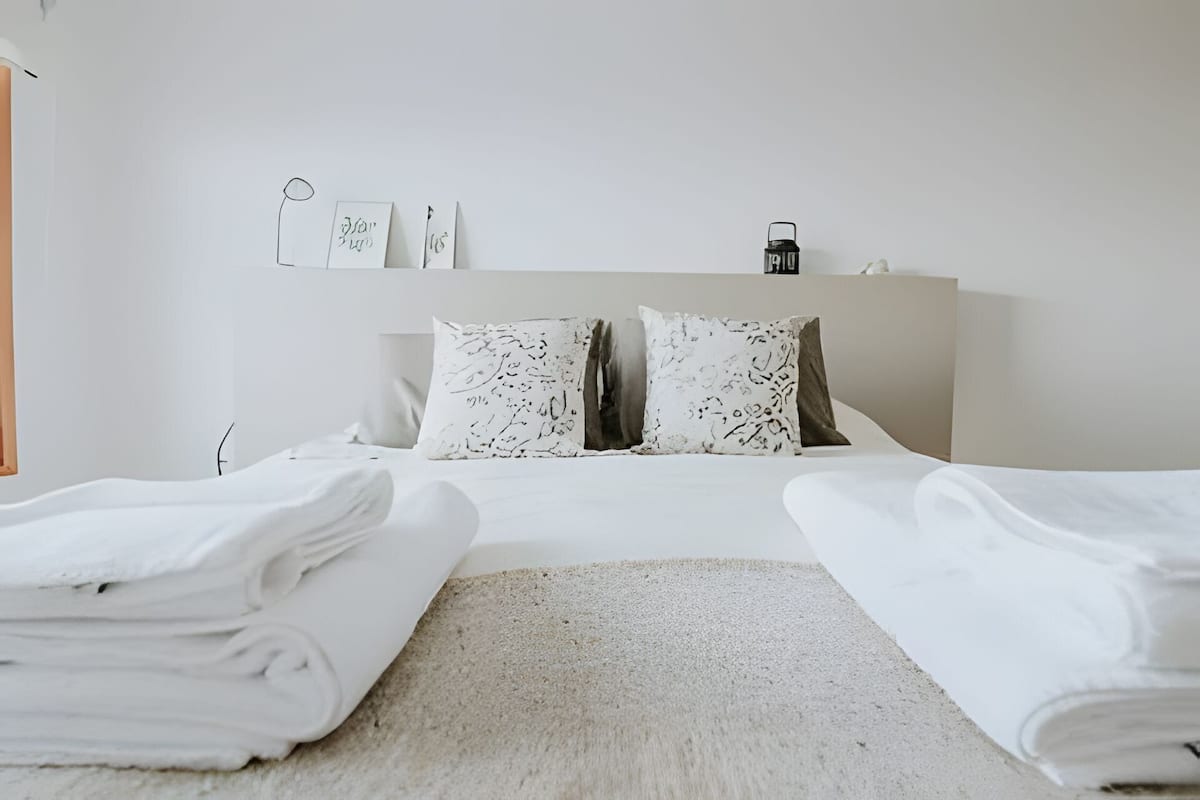 Un lit bien fait, avec du linge de lit blanc, deux oreillers aux motifs abstraits et une table de chevet avec une lampe.