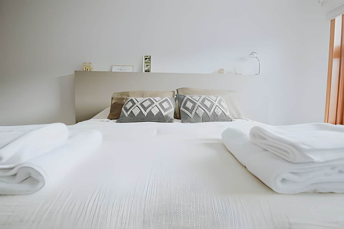 Un lit bien fait, avec du linge de lit blanc et deux oreillers à motifs géométriques.