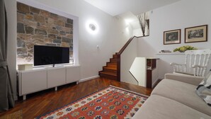 A living room with a stone fireplace, a TV mounted on a white cabinet, a wooden staircase, and a beige sofa. - Casa Marlò Centro Storico (Cividale del Friuli)