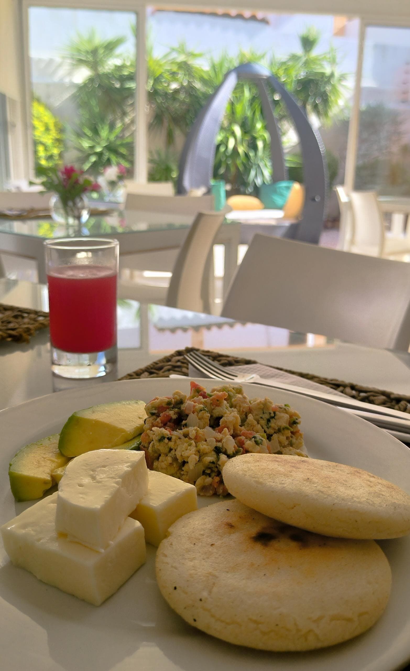 A breakfast plate with scrambled eggs, avocado slices, cheese, and arepas, with a glass of red juice on the table.