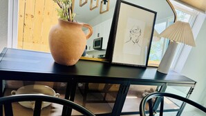 A modern dining area with a glass table, black chairs, a ceramic pot with plants, a framed sketch, and a lamp.