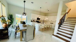 A modern kitchen with white cabinets, a central island with bar stools, a dining table with chairs, and a staircase with wooden steps.
