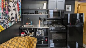 Une cuisine compacte équipée d’un four à micro-ondes, d’une cuisinière et d’un réfrigérateur. On y trouve un évier avec un robinet, et des étagères au-dessus du plan de travail où sont rangés la vaisselle et les verres. Une œuvre d’art colorée est accrochée au mur, à gauche.