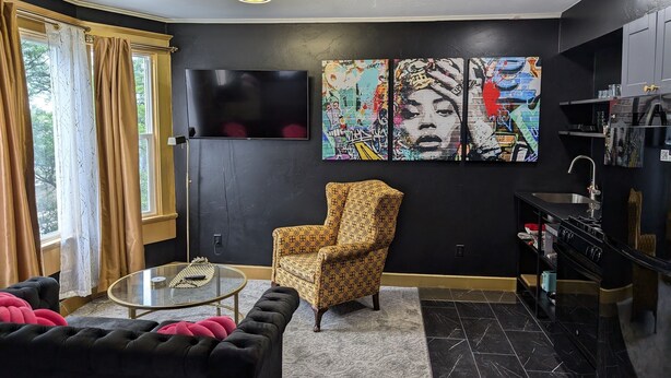 Un salon moderne avec un canapé noir, un fauteuil à motifs, une table basse en verre et un téléviseur fixé au mur.