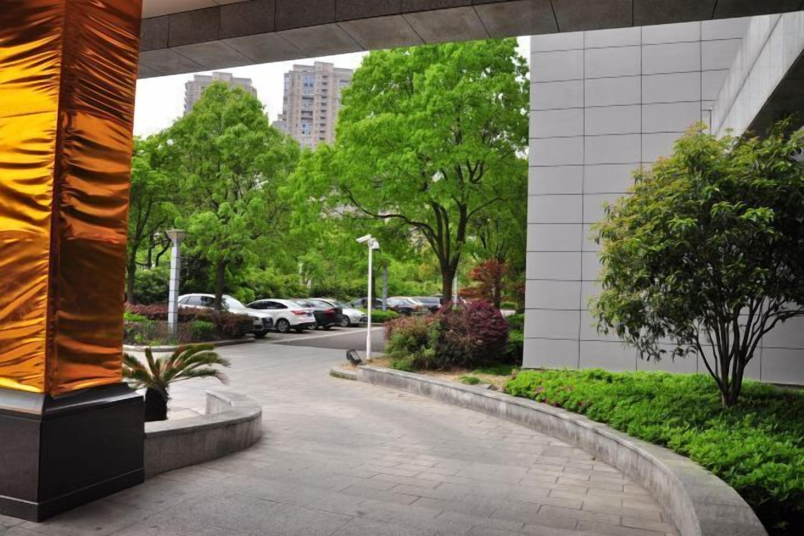 A modern building entrance with a curved pathway, surrounded by lush greenery and parked cars.