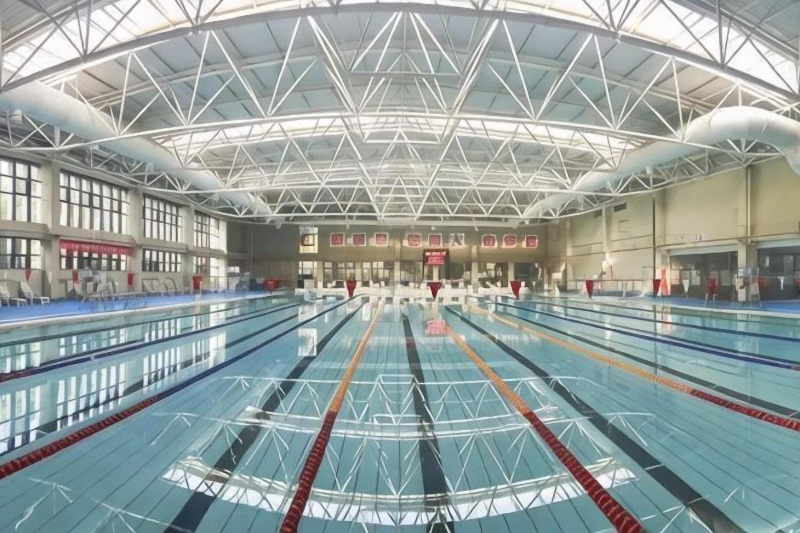 An indoor swimming pool with multiple lanes, a high ceiling with a geometric design, and a large window on the left side.