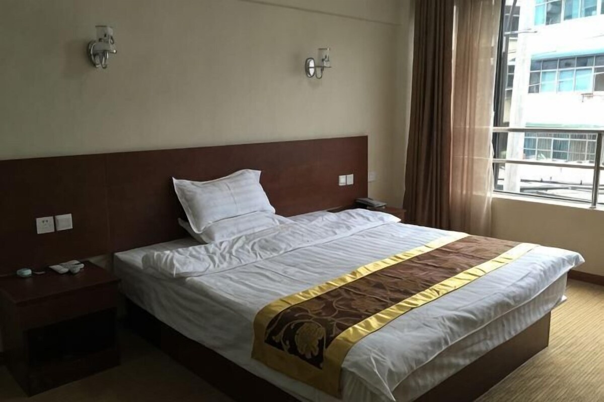 A hotel room with a large bed, bedside tables, a nightstand, a window with a view of buildings, and wall-mounted lights.