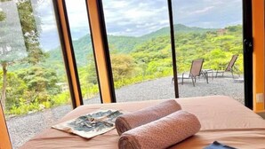Habitació d'hotel amb un llit, una tovallola i vista a un paisatge verd i exuberant a través de grans finestres.