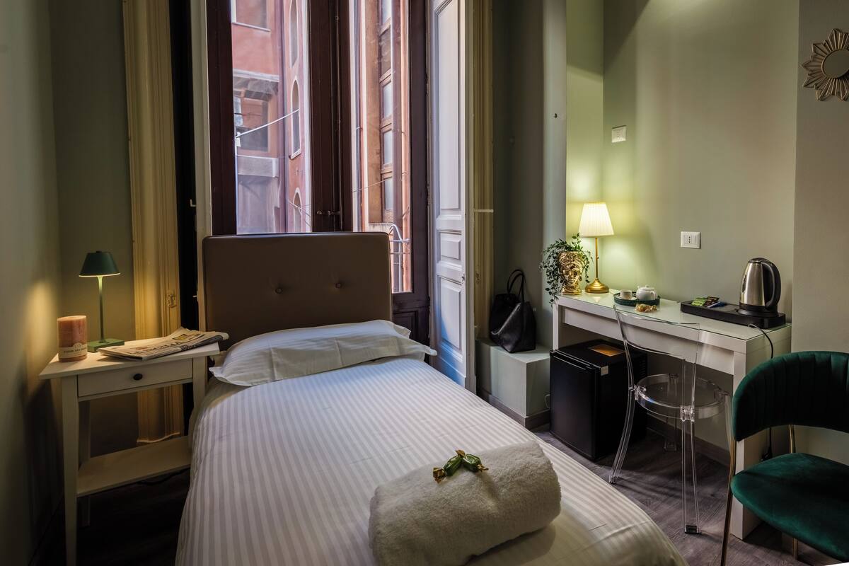 Une chambre d’hôtel équipée d’un lit, d’un bureau, d’une chaise, d’une lampe et d’une fenêtre donnant sur des immeubles.