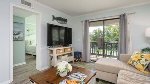 A living room with a sofa, a coffee table with magazines, a TV, and a balcony with a view of greenery. - Tennis Villa F20 - Close to Beach (Hilton Head Island)