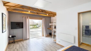 A modern hotel room with a wooden ceiling, a large window with curtains, a flat-screen TV, a bed with a blue blanket, and a white radiator.