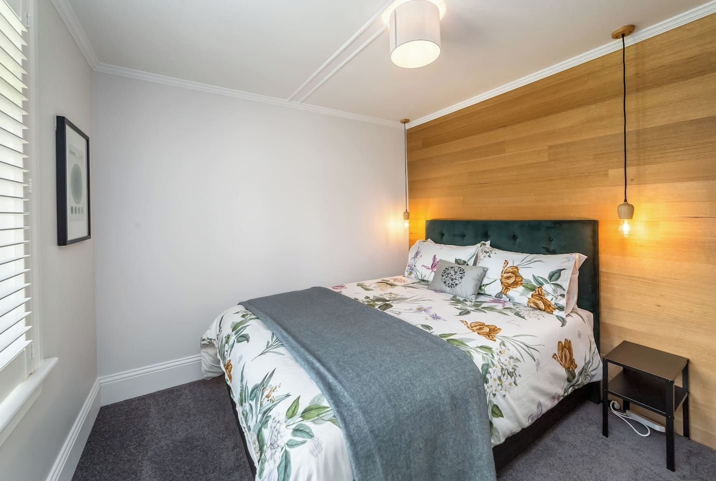 A bedroom with a bed, a bedside table, a wall-mounted light, and a window with blinds.
