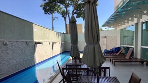 Un área de piscina al aire libre con mesas y sillas de madera, sombrillas y sillones de descanso.