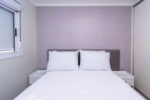 Una cama bien hecha con sábanas y almohadas blancas, un cabecero gris y una ventana con persianas.