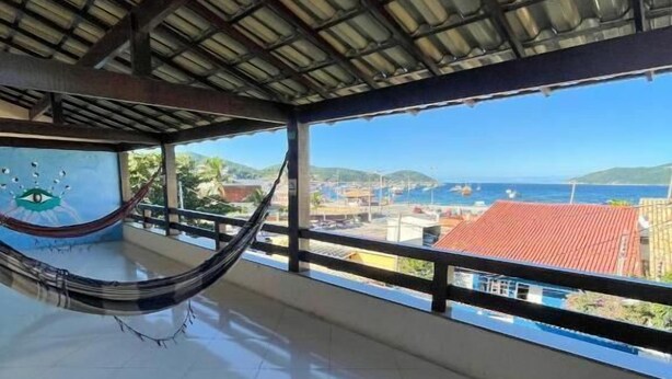 A room with a hammock, a tiled floor, a view of the sea, and a building with a red roof. - Enseada Hostel (Arraial do Cabo)