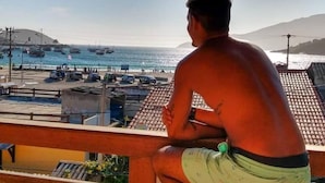 A person sitting on a balcony overlooking a beach with boats and a clear sky. - Enseada Hostel (Arraial do Cabo)