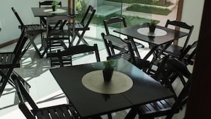 A dining area with multiple tables and chairs, a glass door leading outside, and a view of a garden. - Enseada Hostel (Arraial do Cabo)