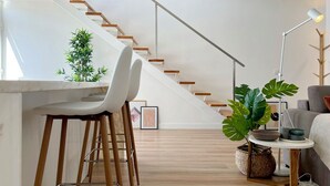 A modern living space with a staircase, a dining area with bar stools, and a side table with a lamp and decorative items.
