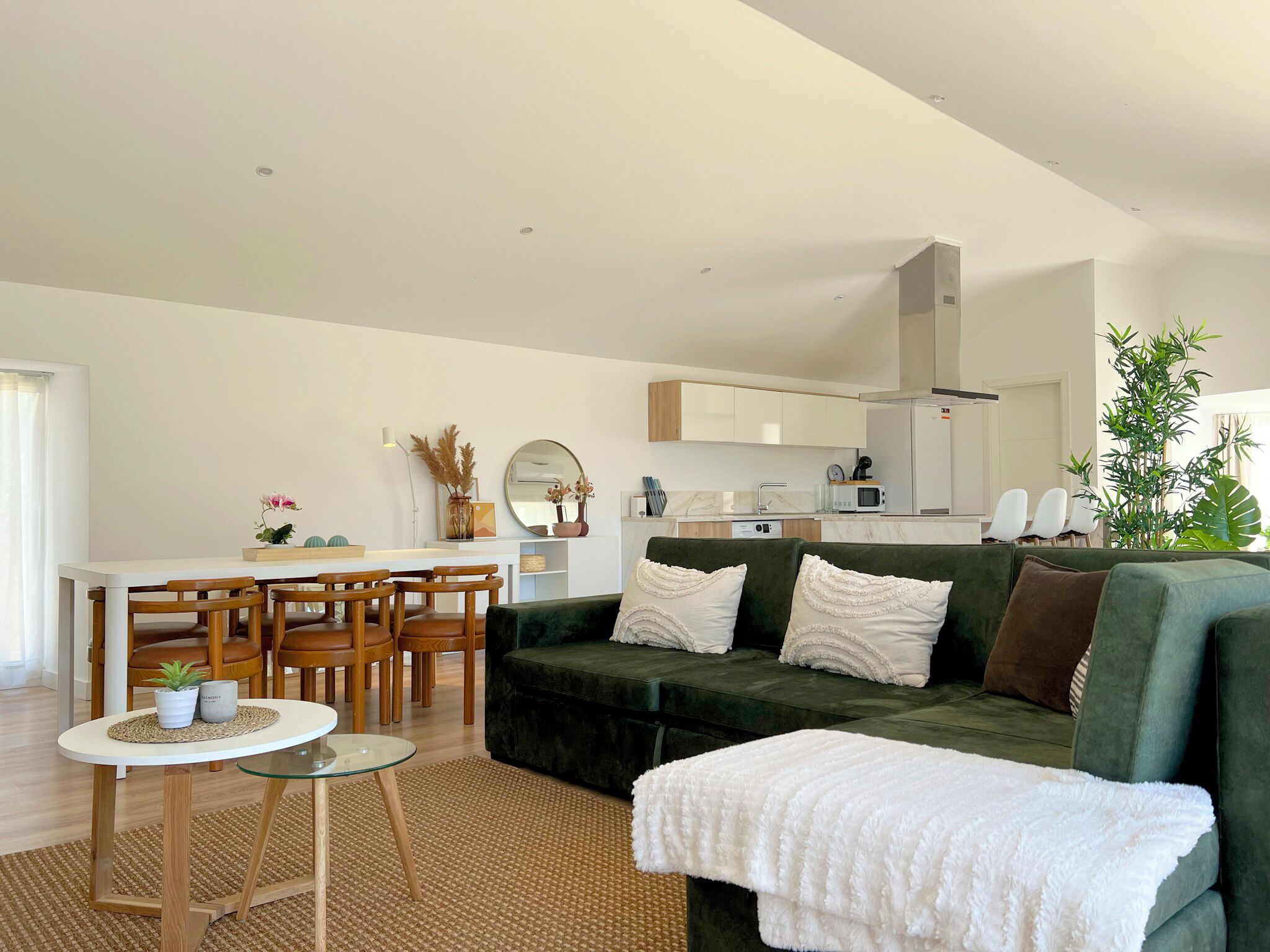 A modern living room with a green sofa, a dining area with wooden chairs, a kitchen with white cabinets, and a large mirror above the counter.