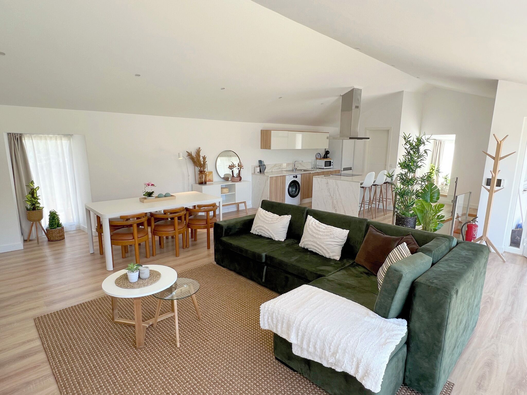 A modern living room with a green sofa, wooden dining table, and chairs. There is a kitchen area with white cabinets and a washing machine.