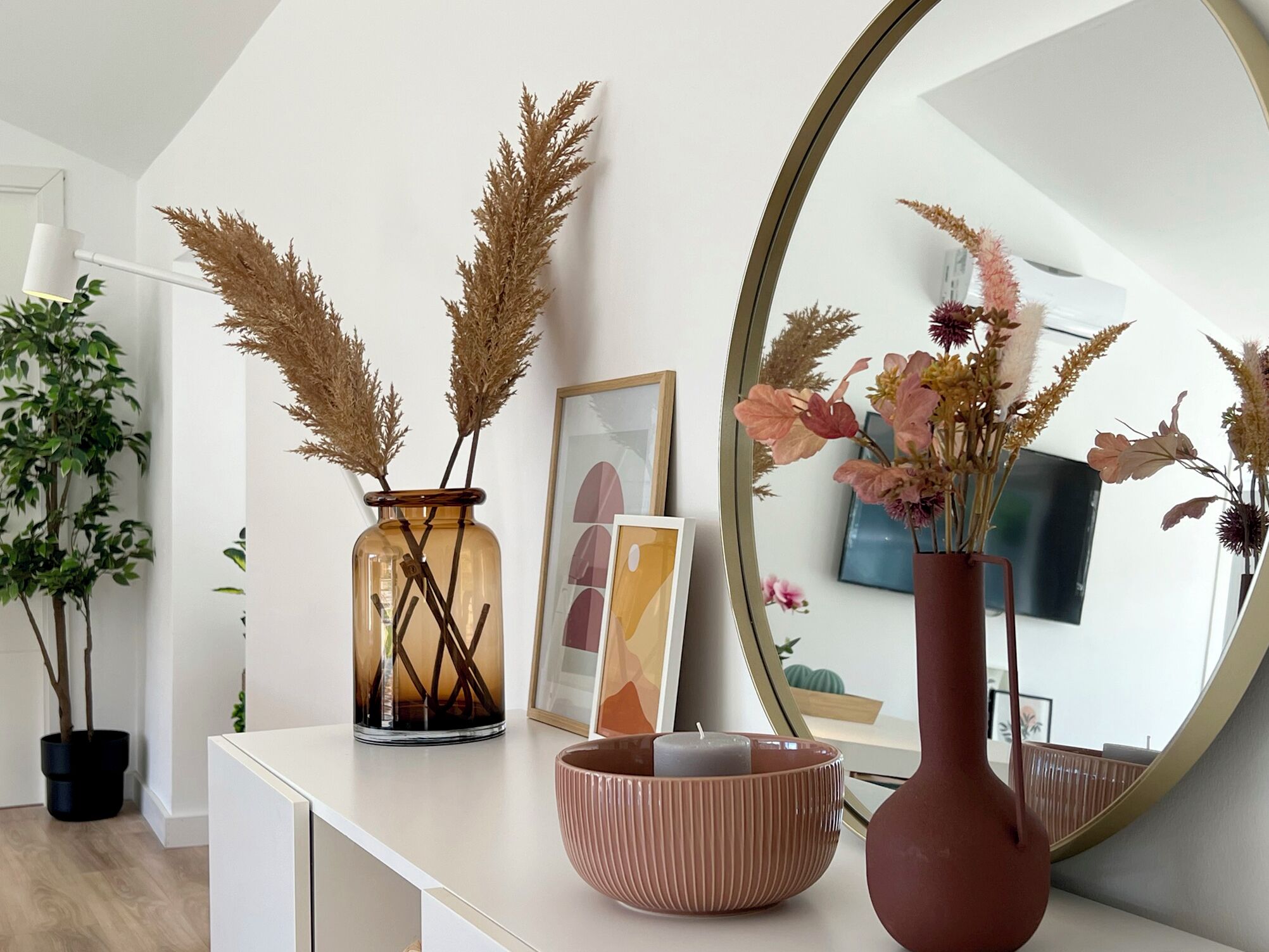 A white console table with a mirror, a vase with dried pampas grass, a framed picture, and a pink bowl with candles.