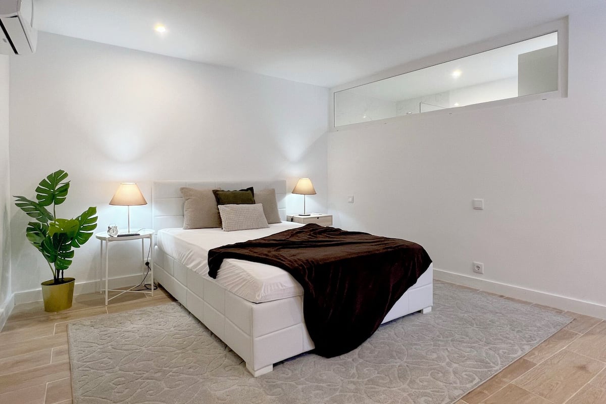 A modern bedroom with a white bed, two bedside lamps, a large mirror, and a potted plant.