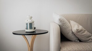 Une lanterne blanche est posée sur une petite table ronde aux pieds en bois, à côté d’un canapé beige agrémenté de deux coussins.