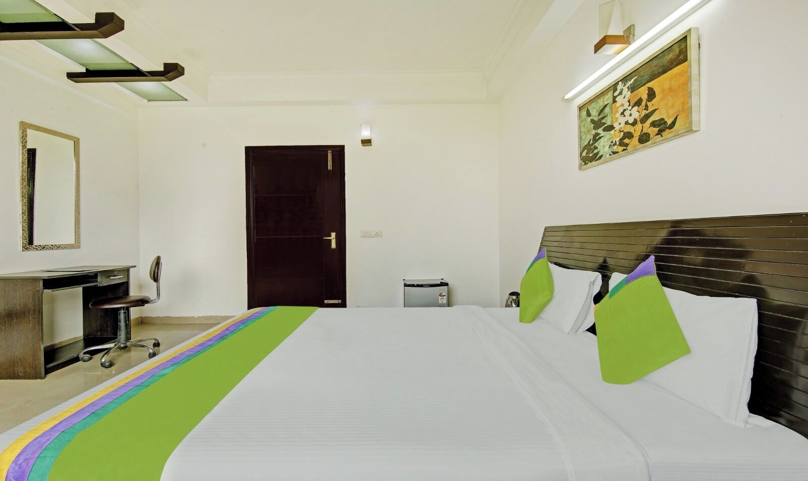 A hotel room with a bed, a desk, a chair, a door, and a wall-mounted light fixture.