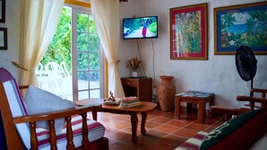 A cozy living room with a television, two paintings, a fan, and a table with books and a vase. - Charming 3-bedroom Gingerbread Style Villa in Bon Accord, Tobago (Bon Accord)