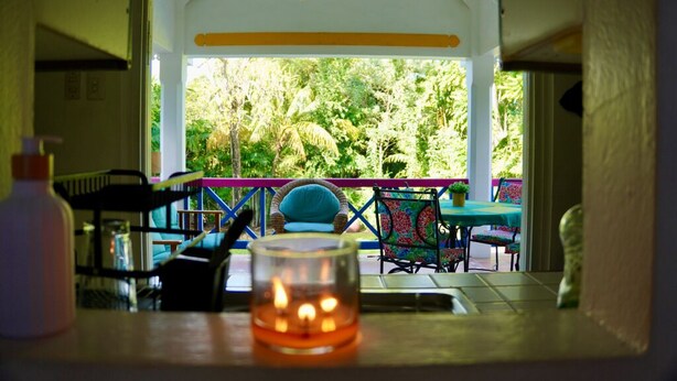 A room with a view of a lush garden, a table with chairs, and a lit candle on the table. - Charming 3-bedroom Gingerbread Style Villa in Bon Accord, Tobago (Bon Accord)