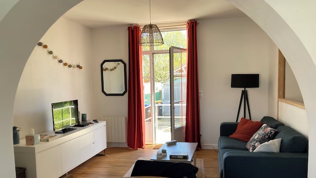 A cozy living room with a wooden table, a sofa, a TV, and a large window with curtains. - Pretty, quiet vacation home with wooded garden  (Perigny)