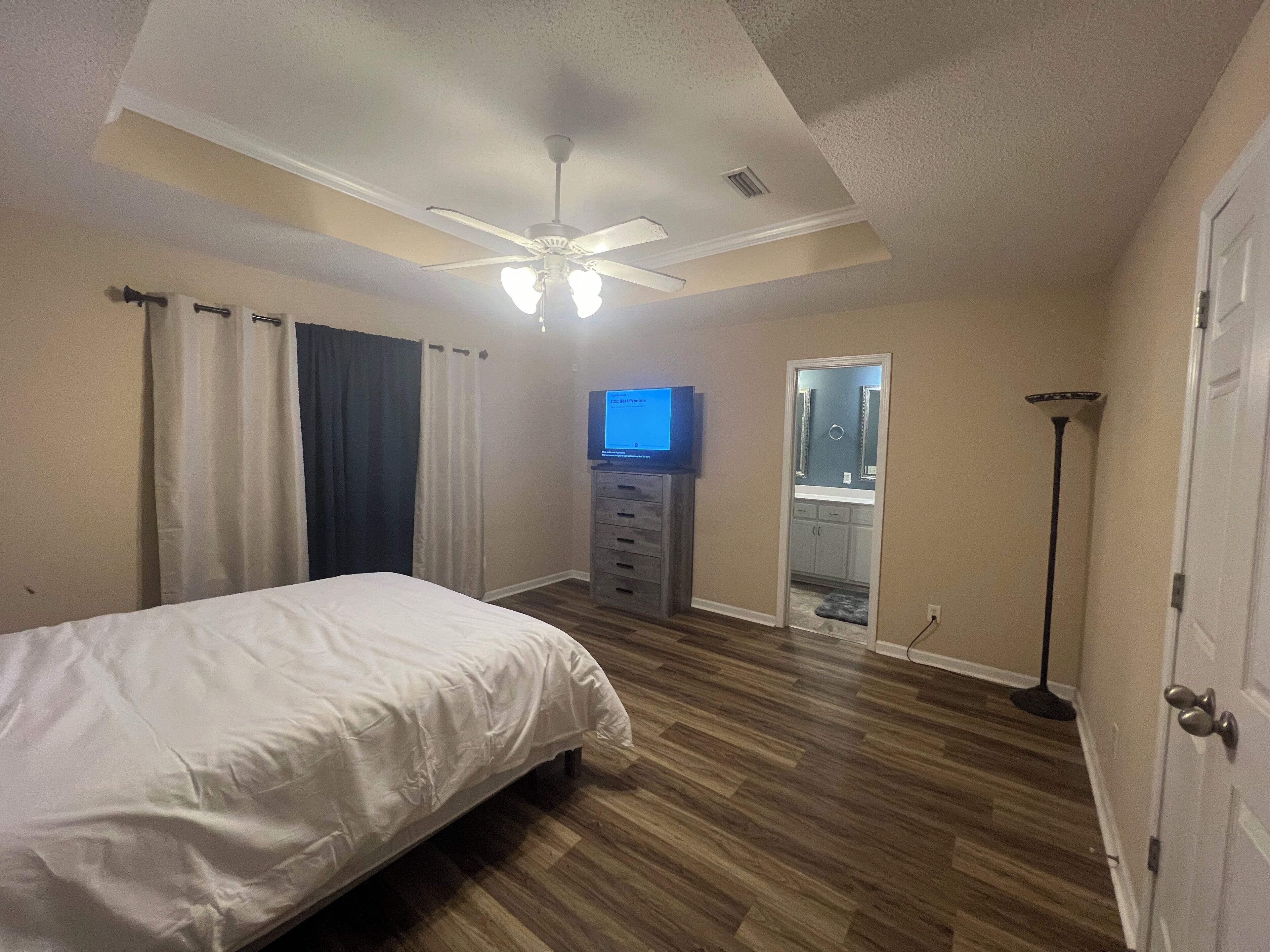 A bedroom with a bed, a ceiling fan, a television, a mirror, and a door.