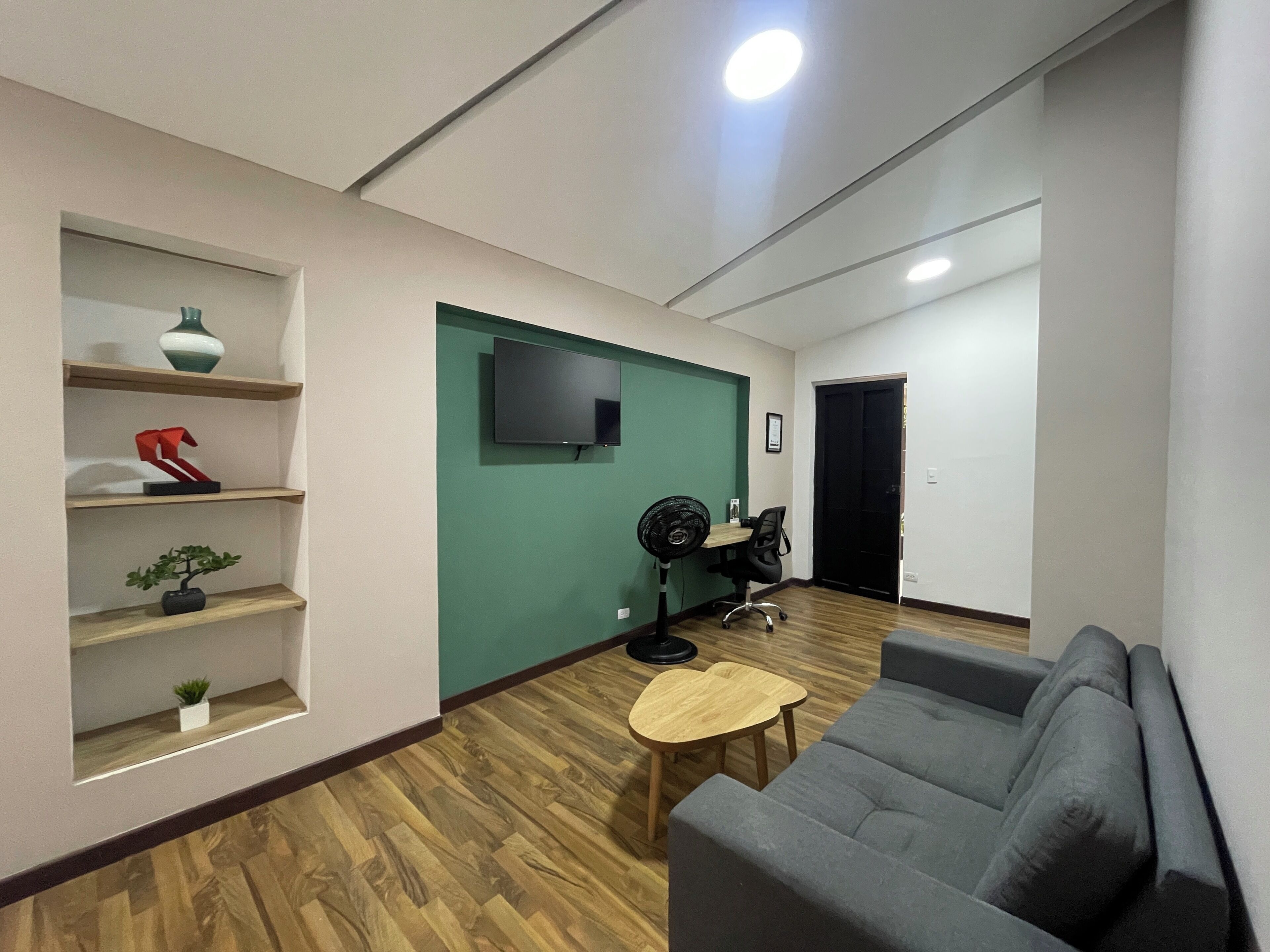 A modern living room with a grey sofa, wooden flooring, a green accent wall, a mounted TV, and a small table with a fan.
