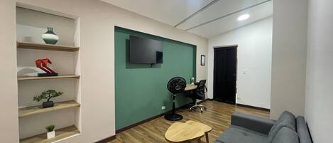 A modern living room with a grey sofa, wooden flooring, a green accent wall, a mounted TV, and a small table with a fan.
