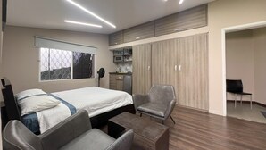 Une chambre moderne comprenant un lit, deux fauteuils, une petite table, une armoire et un coin cuisine.