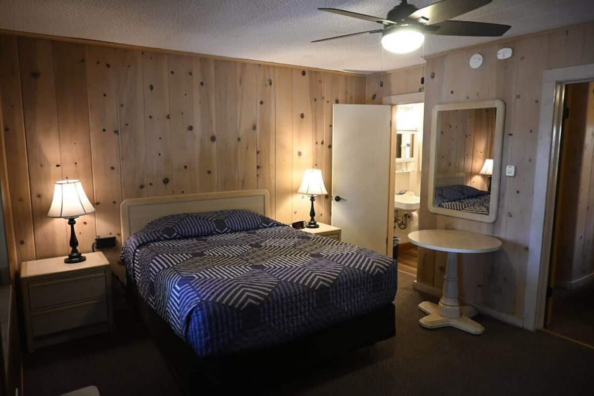 Un dormitorio con una cama, dos mesitas de noche con lámparas, un ventilador de techo, un espejo y una pequeña mesa redonda.