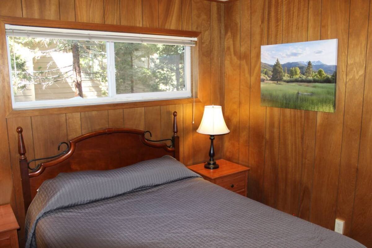 Una habitación con paneles de madera, una cama, una mesita de noche con lámpara, una ventana con vistas a árboles y un cuadro de paisaje en la pared.