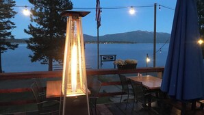 A deck with a table and chairs, a tall heater, an American flag, and a view of a lake and mountains. - Plumas Pines Resort (Canyondam)